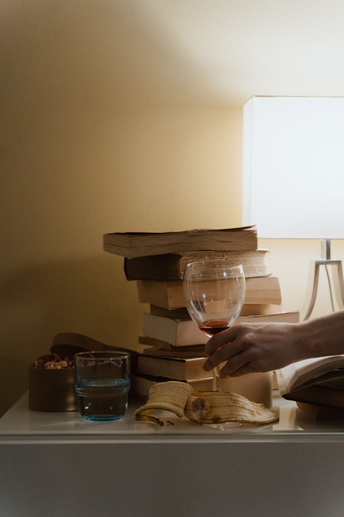 Une main tient un verre de vin rouge près d'une lampe et d'une pile de livres, dans la lumière dorée d'un soir à la maison