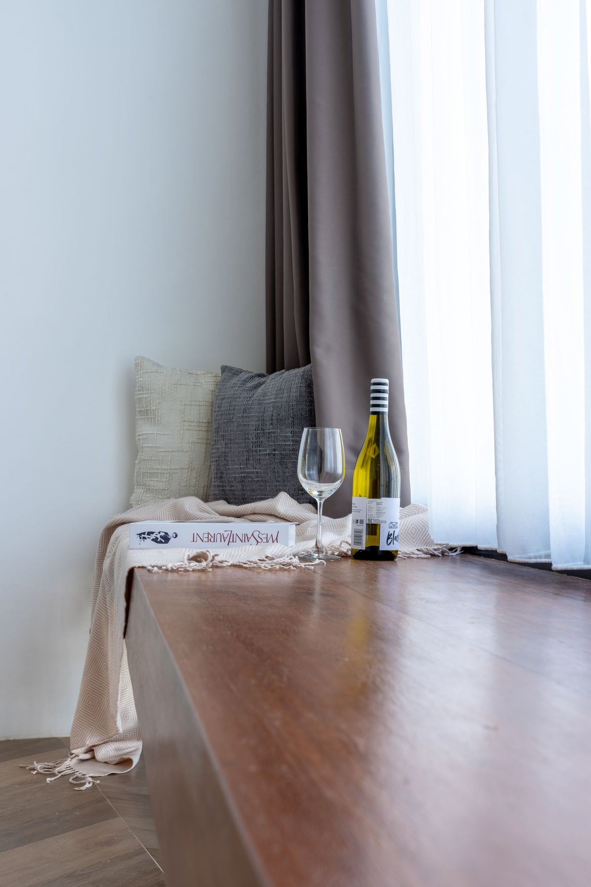 Une bouteille de vin et un verre posés près d'une fenêtre ensoleillée, avec des coussins et un rideau